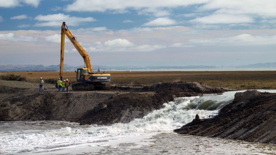 Construction equipment moving away after breaching a levee: water is pouring through the breach and flooding a lower area.