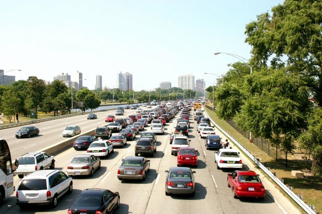 Image shows a line of cars driving down a highway. The side of the highway is lined by vegetation and a fence. A city is shown off in the distance.