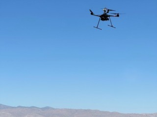 A drone flies above a landscape with three researchers standing in the background.