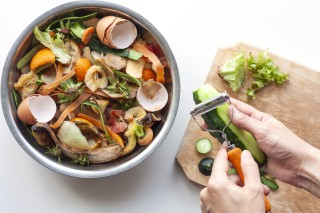 someone peeling a cucumber and putting the peels in the compost bin right next to the cutting board that has eggshells and other fruit and vegetable scraps in it.