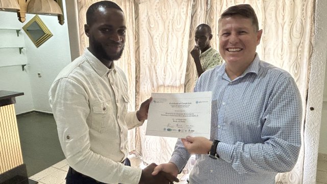 Man wearing white dress shirt smiling at camera and receiving a certificate of completion from man wearing sky blue dress shirt.