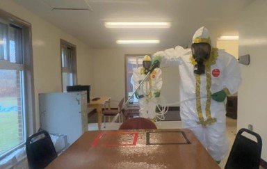 Two responders in a Tyvek suit stand in a room with one of them spraying decontaminant onto a wooden table with a red and black square outline.