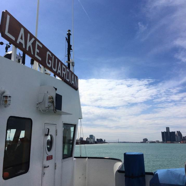 The Lake Guardian boat with The Ambassador Bridge in the background, ona partially cloudy day. The words Lake Guardian visible on the side of the boat.