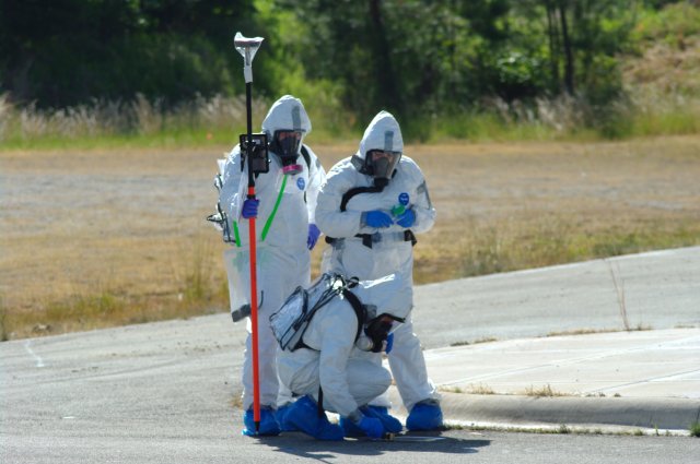 Three team members in tyvek suits with one person taking a sample of the road surface while the other two stand over them with a pole that holds an ipad and GPS unit.