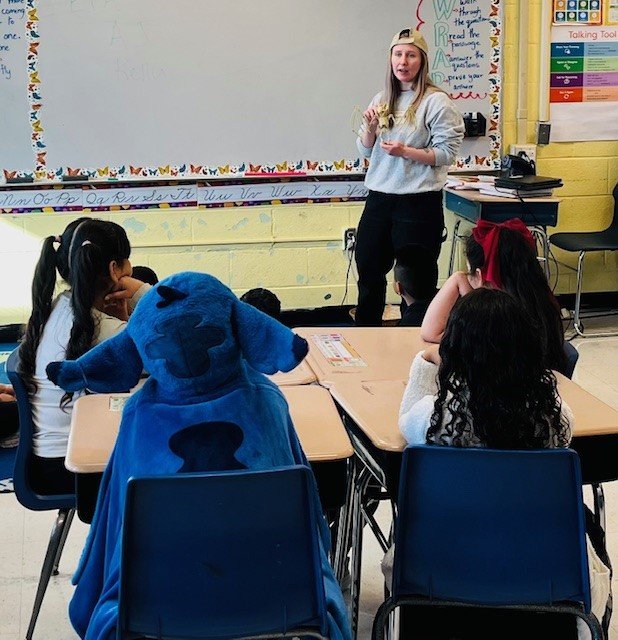 AJ stands at the front of a classroom and kids at a table look on