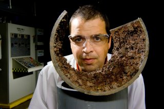 EPA researcher in white lab coat and safety goggles looks over a cross section of old, corroded water pipe. 