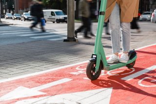 Shows a person on an electric scooter on a dedicated cycling lane in the foreground, and pedestrians on a crosswalk in the background.