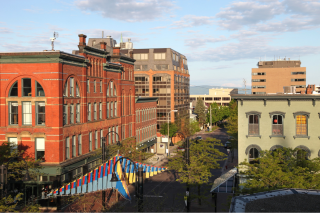 Elevated view of downtown Burlington Vermont office buildings along a street. A larger orange colored building with windows is on the left-hand side of the screen with a lower cream building on the right with a street in between them. Some trees line the street. 