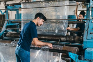 Two people working with a machine that produces a large roll of plastic wrap.