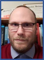 A closeup of a man with glasses, short brown hair, and a short beard. He is wearing a blue collared shirt.