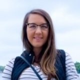 A woman with long brown hair, glasses, and a striped shirt standing outside.