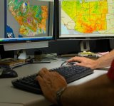 Image of two monitors with images of maps and a person's hands typing on a keyboard and using a mouse.