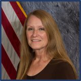 A woman with light skin, long auburn hair, and a dark shirt. She is standing in front of a gray background and American flag.